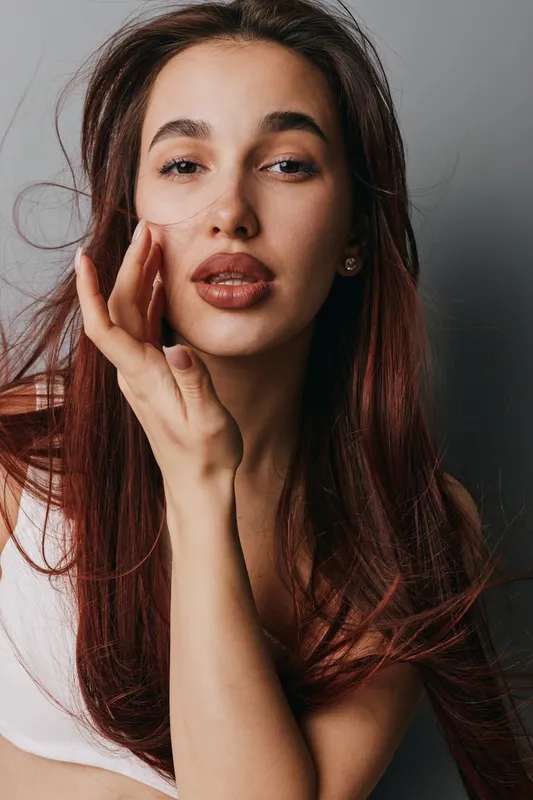 Woman with long red hair and natural makeup posing with hand near face for beauty portrait and skin care concept in studio with soft light and neutral background.