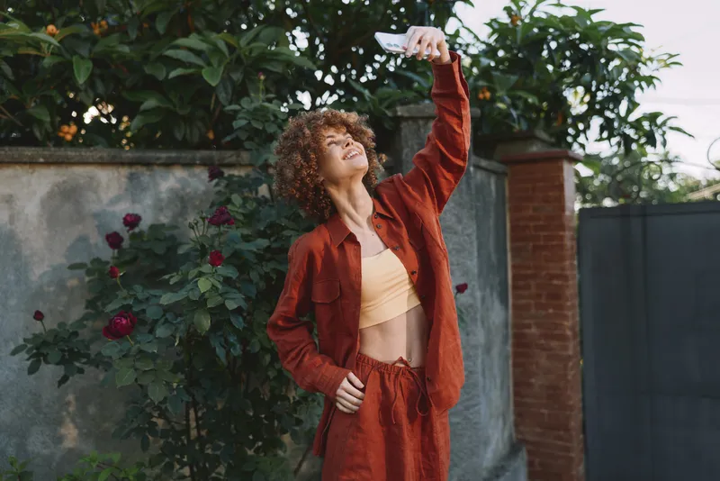 Smiling woman with curly hair outdoors in a red outfit, capturing a lively lifestyle moment, garden backdrop, candid portrait, warm natural light, emphasizing self expression and confidence