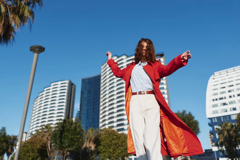Brunette woman in a red trench coat and white shirt stands outdoors in a city plaza on a sunny day, arms raised, beige trousers, tall buildings framing blue sky.