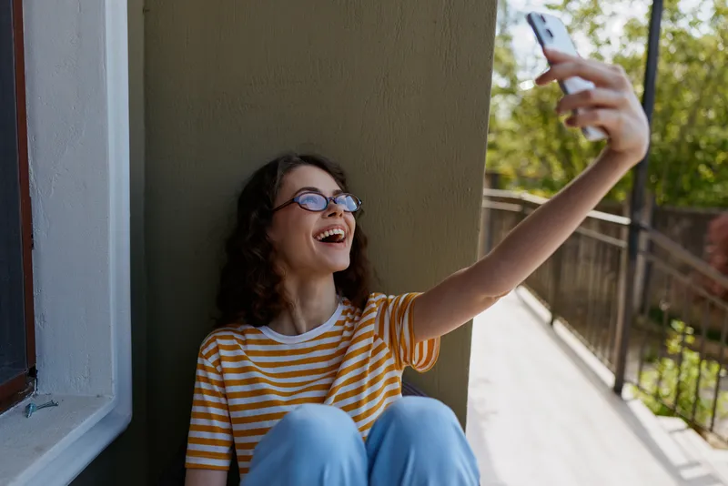 A young woman with curly hair and glasses sits beside a balcony wall and laughs while taking a selfie in a yellow striped tshirt, capturing a bright casual moment.