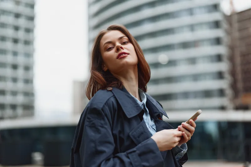 A young woman with brown hair in a navy jacket looks up from her phone amid a modern cityscape, capturing a moment of urban exploration and contemplation.