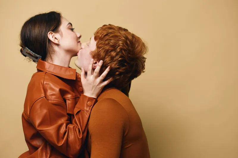Couple in love with affectionate embrace and kiss, young man and woman wearing casual brown clothes with warm beige studio background and emotional connection.