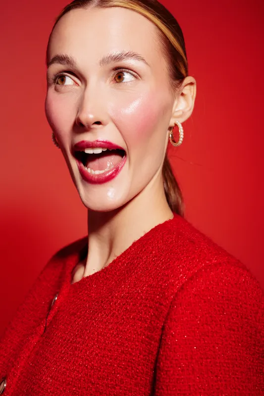 Young woman with shiny skin and red lips wearing red sweater and hoop earrings looks surprised on red background. Concept of beauty, fashion, and vibrant emotions.