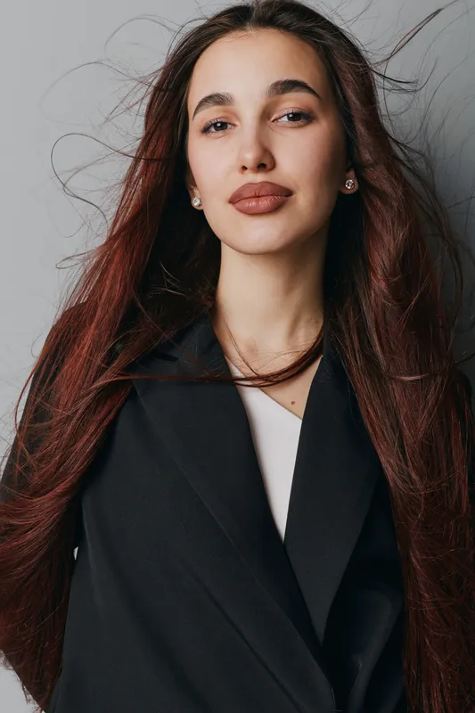 Woman with long hair wearing black coat posing against gray wall, looking confident and stylish with natural makeup and earrings in portrait indoors with soft lighting.