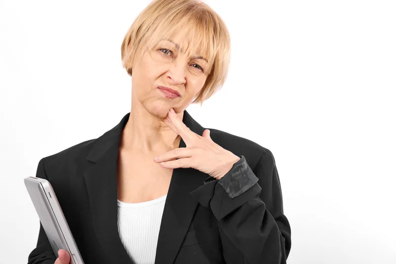 Senior woman portrait in studio light, holding smartphone, touching chin, thinking concerned worried expression, blond hair, black jacket, white background, copy space, calm
