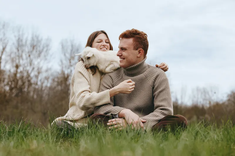A smiling couple sits on a grassy field in knit sweaters, a fluffy dog nuzzles them, capturing a warm moment of affection and natural lifestyle outdoors.