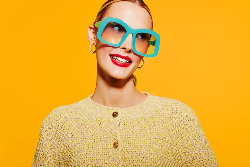 Smiling woman with red lipstick and large turquoise sunglasses wears yellow knitted sweater on bright orange background. Concept of fashion, style, and cheerful mood in studio.