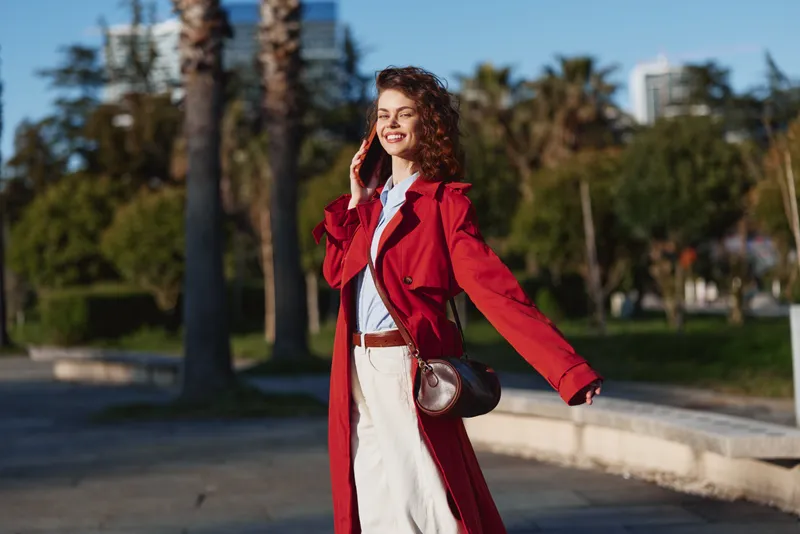 Young woman with curly hair wearing a red coat talks on the phone and smiles while walking outdoors on a sunny day near palm trees.