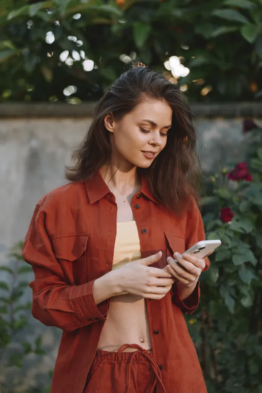 A woman in a rust jacket checks her phone in a sunlit garden, casual fashion mood. A lifestyle portrait highlighting everyday style, confidence, and relaxed moments