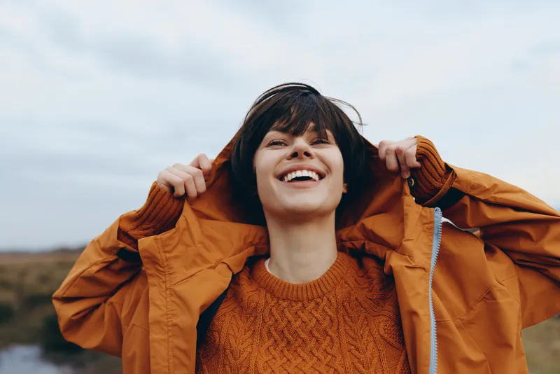 Smiling woman outdoors laughing in the wind, holding a jacket hood, casual sweater and warm clothing, lifestyle moment under cloudy sky, open landscape, fresh breeze, natural light today
