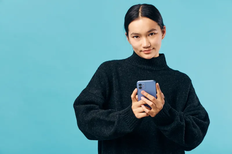 Young woman wearing black sweater holding a smartphone and looking at camera with slight smile. Portrait with blue background and casual modern style for technology use and communication.