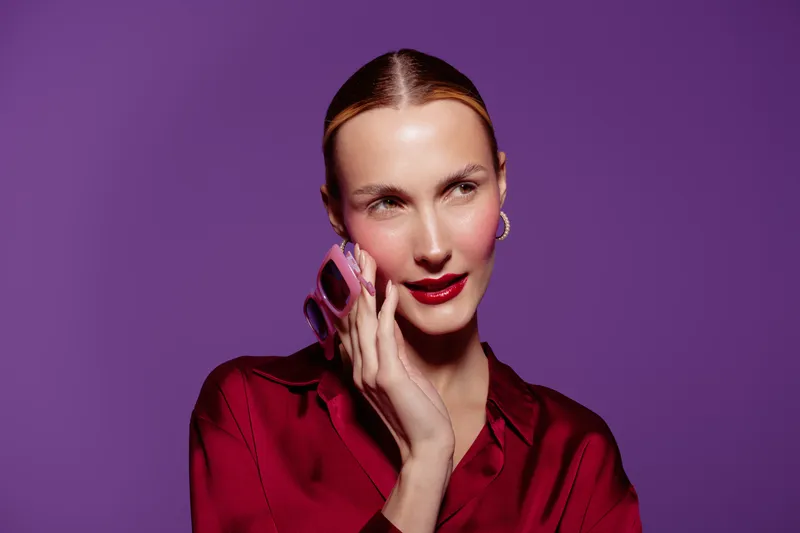Young woman with slicked hair wearing red satin blouse talks on smartphone against purple background. Concept of communication, technology, and modern lifestyle in studio lighting.