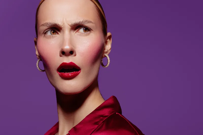 Close-up of surprised woman with red lipstick and earrings wearing red satin blouse on purple background. Concept of emotion, beauty, fashion, and female expression.