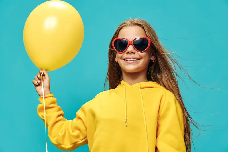 Girl in yellow hoodie holding yellow balloon smiling with heart shaped sunglasses on blue background in studio lighting for celebration and joy youth happiness.