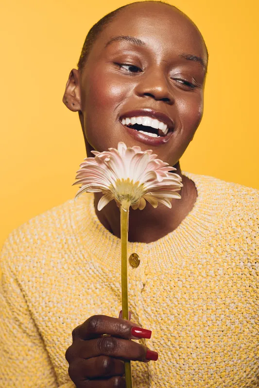 Happy African woman with dark skin holds pink flower and smiles. She wears yellow sweater and poses on yellow background. Concept of joy, beauty, nature, and positive emotions.