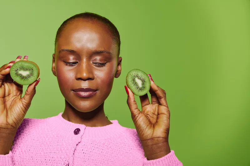 Portrait of dark skinned African woman in pink sweater holding two kiwi halves near face with closed eyes. Concept of healthy lifestyle, natural food, and fresh fruit on green background.