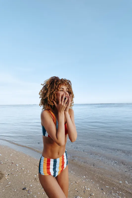 Woman, beach, ocean, swimsuit, hands, face, striped, summer, shoreline, sky, curly haired young woman stands on sandy shore, covers eyes with palms, shy pose, calm waves, soft daylight