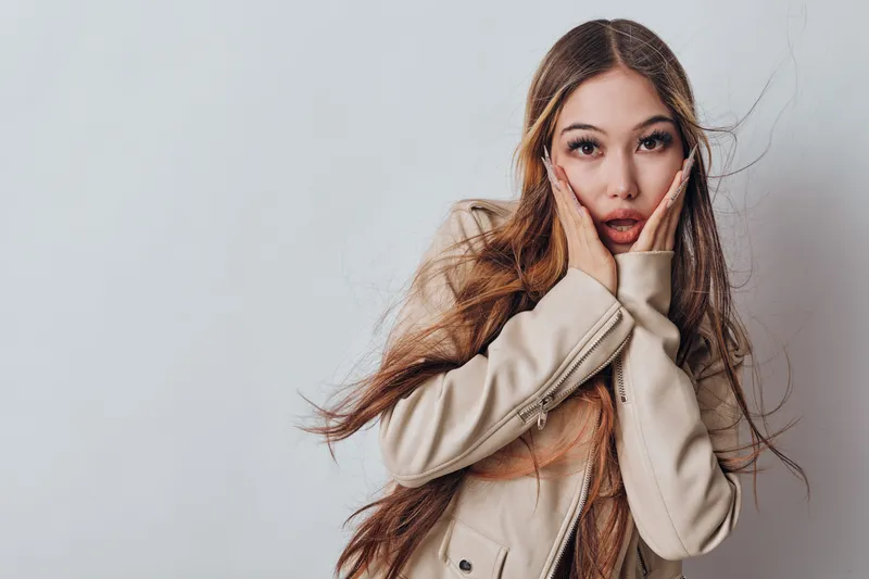 Woman with long hair and casual jacket showing surprised expression and holding face with hands against neutral background with copy space on side.