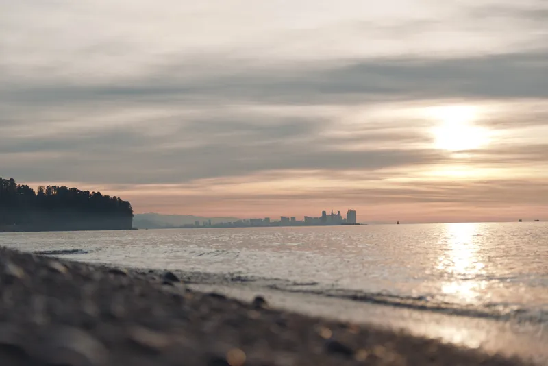 Sunset over a calm sea meets a quiet beach with distant silhouette on the horizon, soft clouds glow, gentle waves, and warm light reflecting on the water and shoreline.