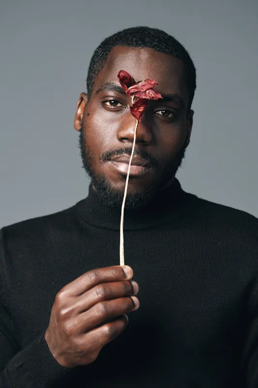 African man with dark skin holding dried flower in front of face in creative conceptual visual style. Portrait of thoughtful black male wearing black turtleneck on gray background.