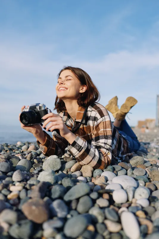 Female photographer lying on a pebble beach with camera, smiling under a clear blue sky, enjoying an outdoor photography moment by the sea.