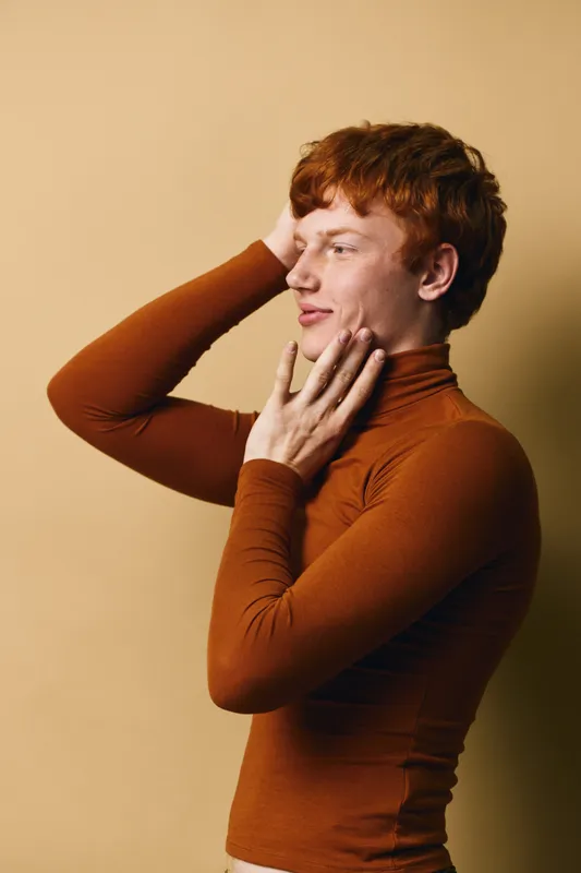 Redhead young man in brown turtleneck posing with hand on face and head in studio with neutral background, profile view for fashion, portrait, and style concept.