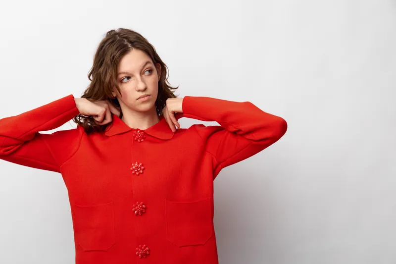 Young woman in red sweater adjusts collar, looking to the side with thoughtful expression. Casual fashion, portrait, and lifestyle concept on plain white background.