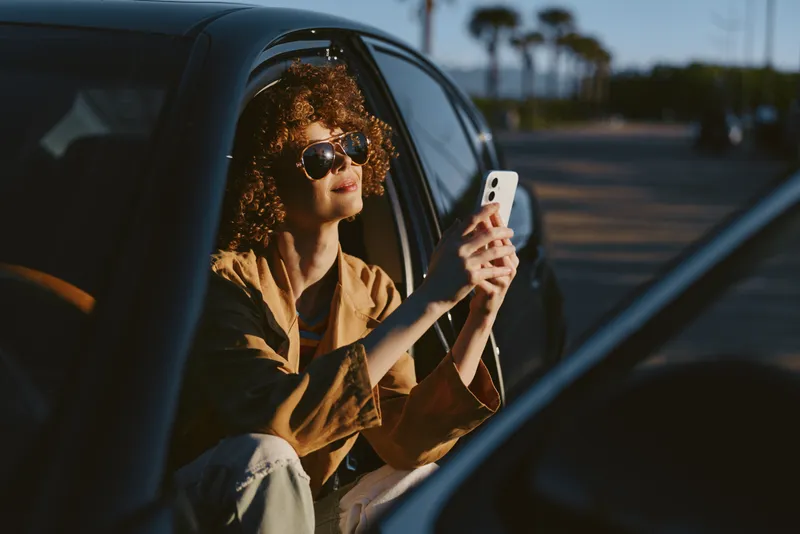 Woman with curly hair sits in a car window, taking a selfie with a smartphone, retro film color tone, casual outfit, relaxed mood, candid lifestyle moment in daylight.