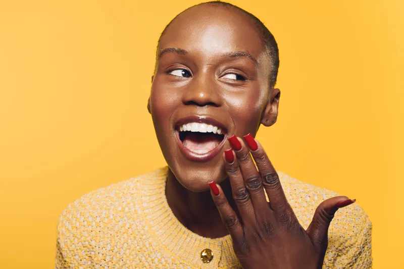 Happy black woman with dark skin smiling and covering mouth with hand showing red nails. Yellow background and cozy sweater create bright, joyful mood. Concept of beauty and happiness.