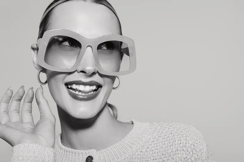 Smiling white woman with fair skin wearing large sunglasses and hoop earrings poses happily in white knitted sweater. Concept for fashion, style, and positive emotions.