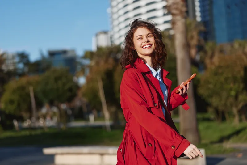 Young woman in a bright red trench coat smiles while walking outdoors in an urban park, sunlight on her face and modern buildings in the background