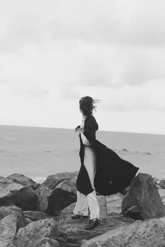 Woman standing on rocky shore, wind blowing her dress as waves crash in the background, black and white scene conveying solitude, movement and contemplation by the sea, a solitary figure framed by