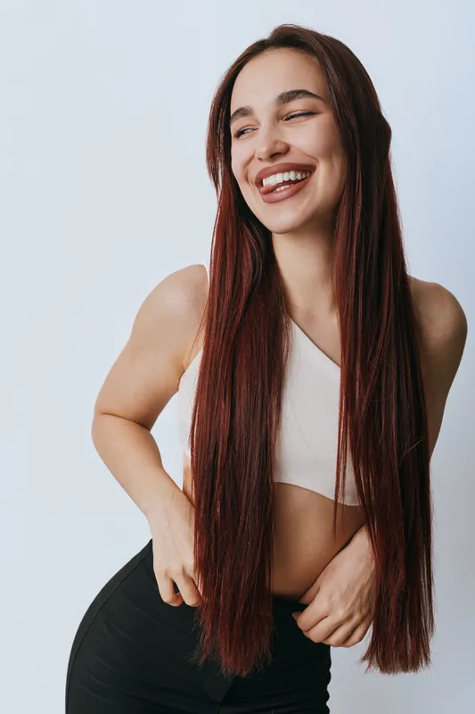 Young woman with long straight hair smiling and posing in casual sportswear against white background. Female model with natural makeup and confident happy expression in studio light.