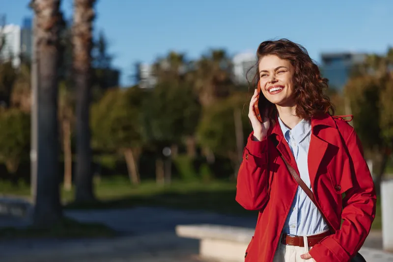 Smiling woman in a red coat talking on a phone outdoors, park setting with trees and blue sky. A candid lifestyle portrait showing fashion, confidence, and cheerful mood.