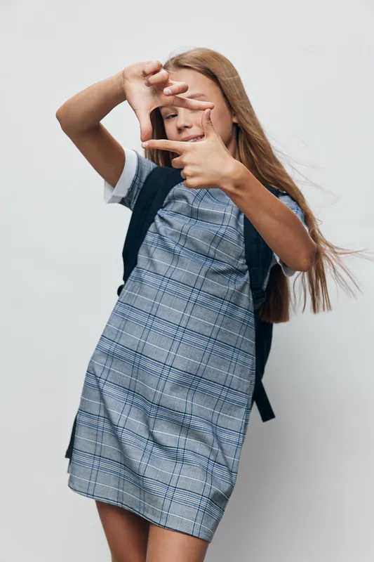Girl with long hair wearing plaid dress and backpack making frame with hands for photo concept isolated on white background. Youth, casual style and creativity in studio portrait.