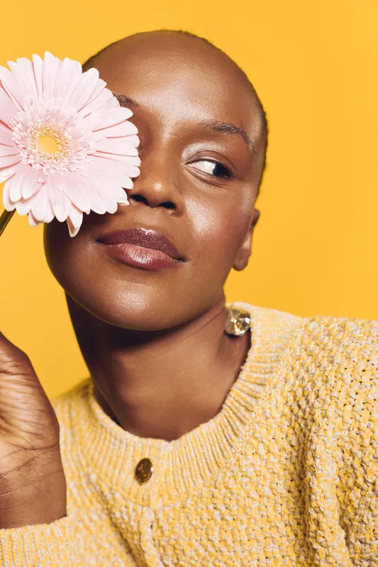 Close-up of a dark skinned African woman holding a pink flower near her face, wearing a yellow knitted sweater. Bright yellow background creates a warm, vibrant mood for beauty and wellness.