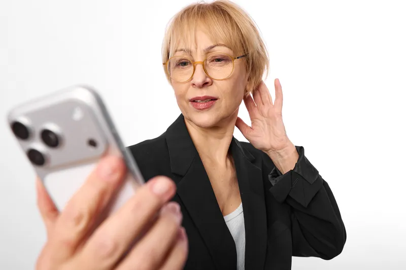 Mature woman with glasses holding smartphone, waving hello at camera, friendly expression, black blazer and white shirt, bright studio background, technology concept