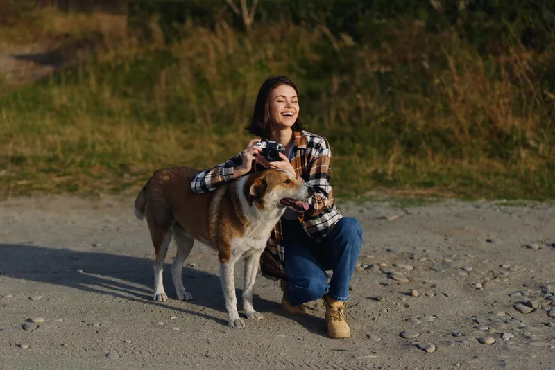 Woman kneeling on a gravel path with a dog, smiling and holding a camera in a sunlit outdoor setting, capturing a moment of companionship and playfulness in nature.