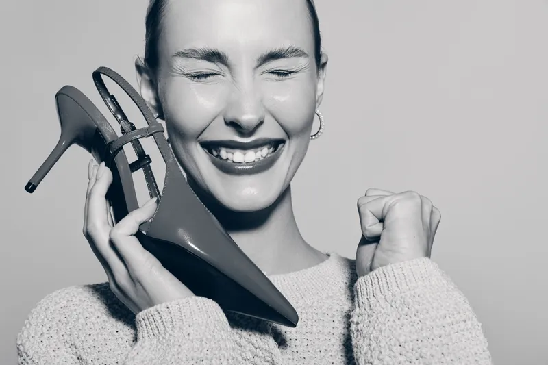 Happy Caucasian woman with light skin holds high heel shoes near face, eyes closed, smiling with joy. Concept of fashion, footwear, style, and positive emotions.
