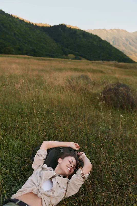 Relaxed woman lying in a grassy field with distant hills and mountains, casual jacket and white top. A natural outdoor lifestyle scene highlighting rest and nature.
