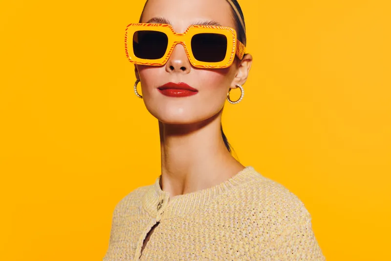 Stylish woman with yellow sunglasses and red lipstick poses confidently against bright yellow background. Fashion portrait with vibrant colors and modern accessories concept.