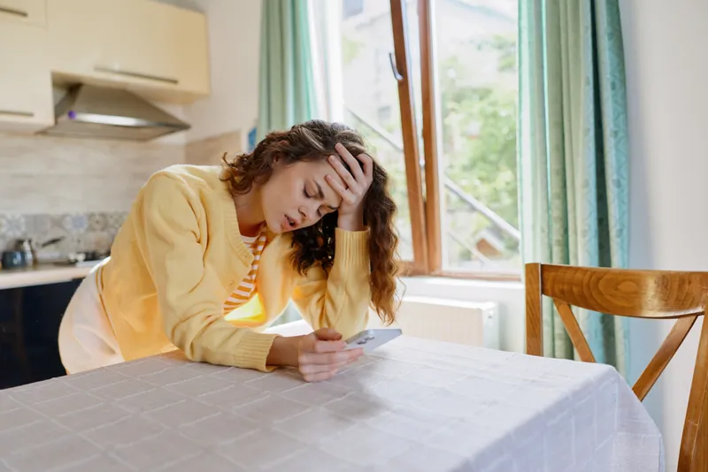 Woman at home sits at a table in a bright kitchen, looking at her phone with a worried expression, supporting head with hand, casual outfit, everyday moment of stress.