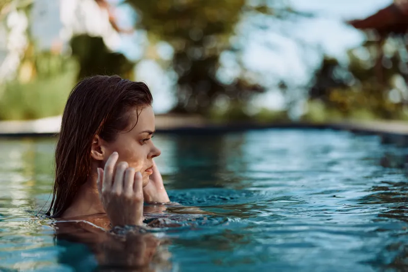 Young woman relaxes in a sunlit pool, fingers resting on her cheeks as gentle water ripples frame a serene moment