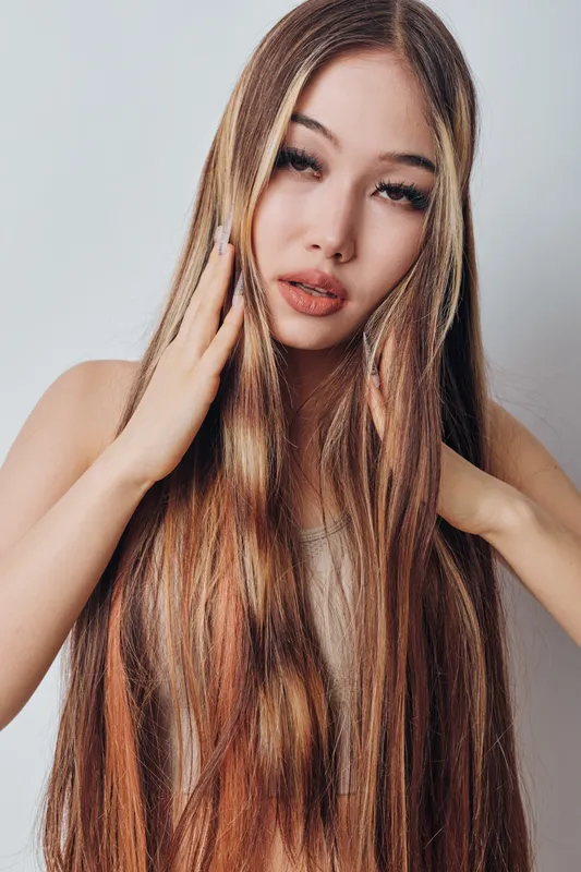 Young woman with long straight hair touching face and hair, looking at camera with soft makeup and neutral background in beauty portrait studio.