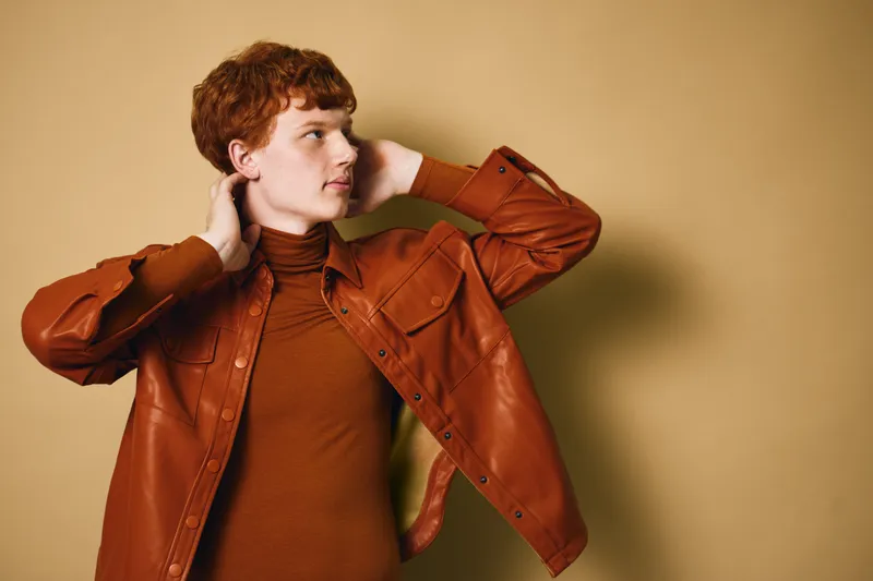 Young man with red hair wearing brown leather jacket and turtleneck posing with hands behind head against beige background. Casual fashion portrait with confident expression.