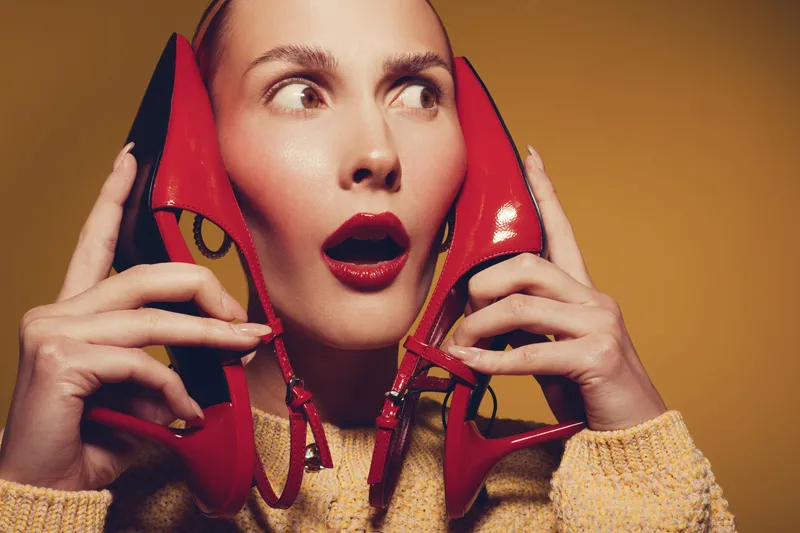 Surprised woman with red lipstick holds red high heel shoes near her face on yellow background. Concept of fashion, style, beauty, and unexpected emotions in studio portrait.