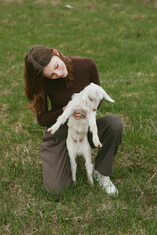 Woman outdoors gently holds a kid goat in a grassy field, capturing a warm lifestyle moment with affection, nature, and animal companionship.
