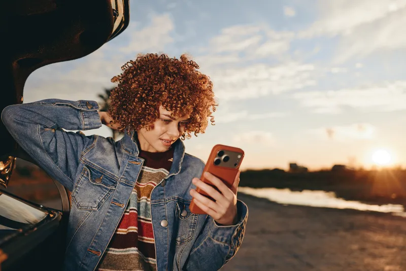 Woman curly hair holding smartphone for selfie in denim jacket, smiling outdoors at sunset with golden light. Lifestyle travel moment in casual style, soft wind and warm sky.