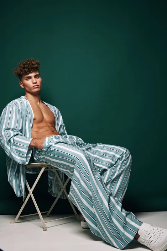 Young man with curly hair wearing striped pajamas sitting on chair against green background, relaxed pose and casual indoor fashion style.