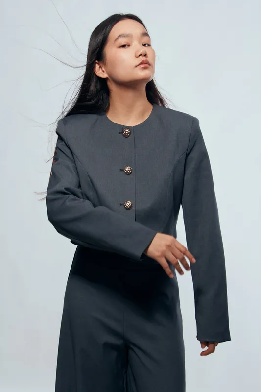 Young woman in modern dark gray suit with embroidered buttons posing confidently in studio. Fashion portrait with long black hair and minimalistic background for style and elegance theme.